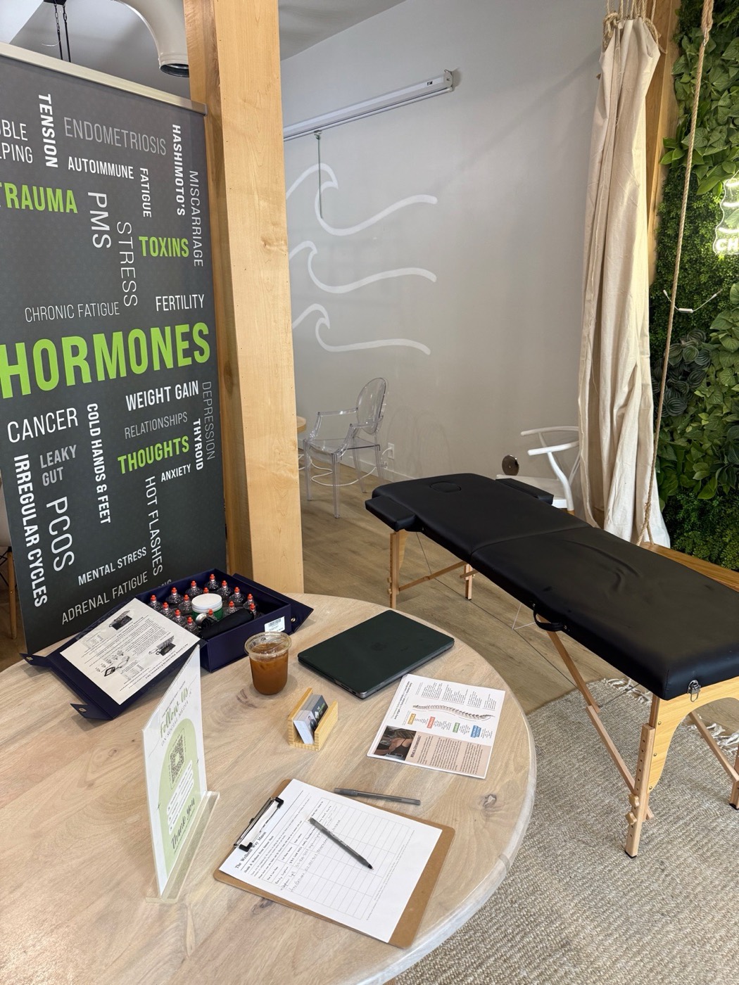 Dr. DeNome's pop-up setup at Chasing Cali Collective — adjustment table, intake clipboard, and a banner listing the symptoms and patterns we see most (hormones, trauma, toxins, thoughts)